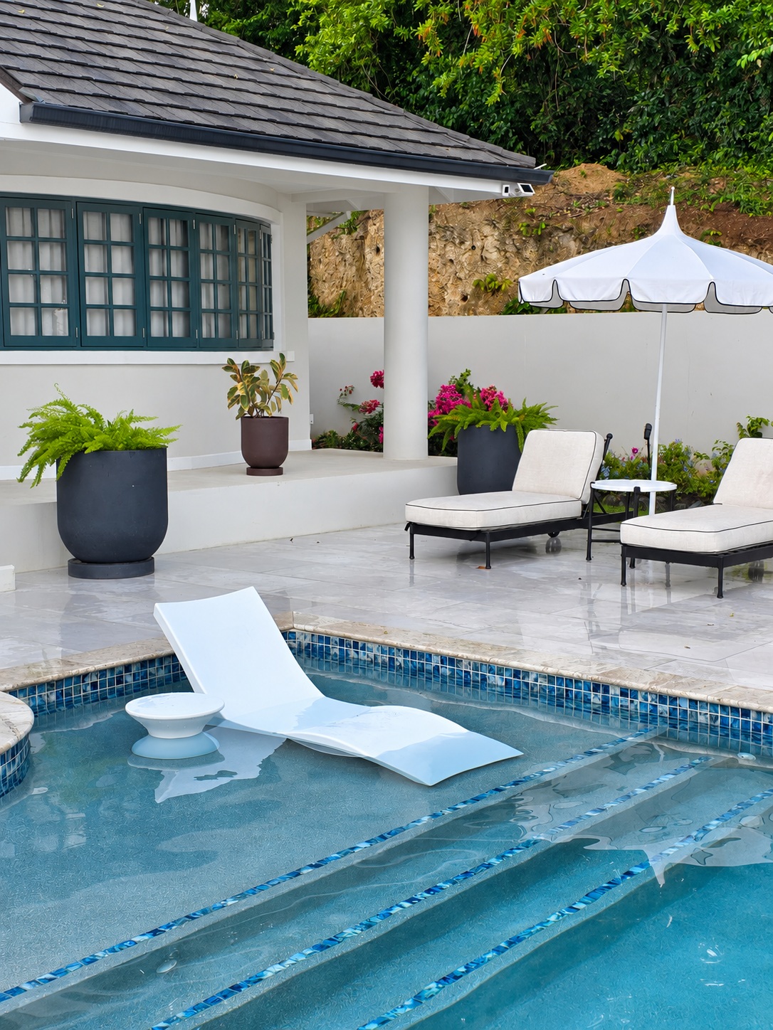 In-pool sun lounger beside the light gray porcelain terrace, loungers and tropical plants